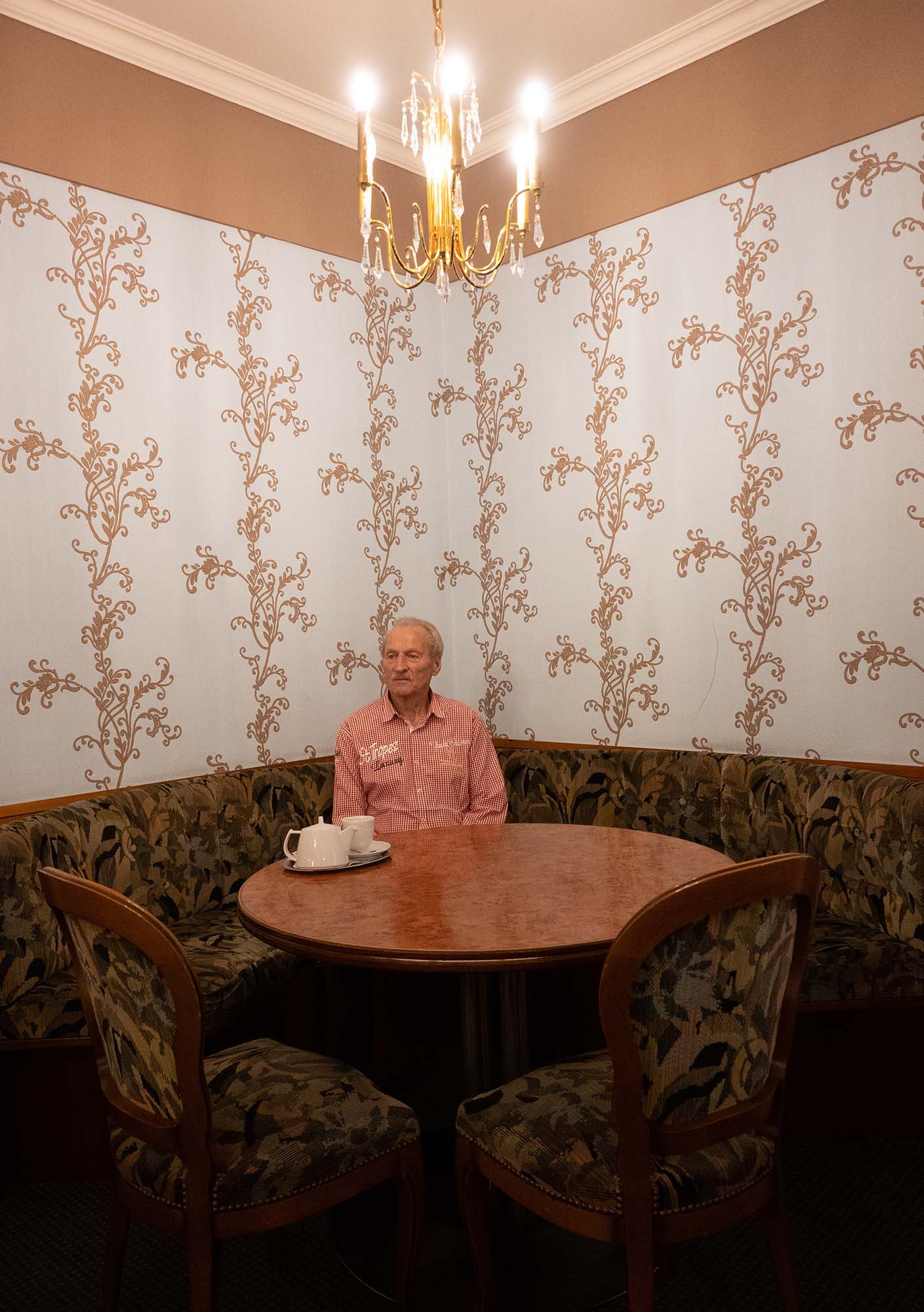 Die Katze sitzt an einem Tisch vor prachtvoller Tapete. Ein goldener Kronleuchter über ihm.