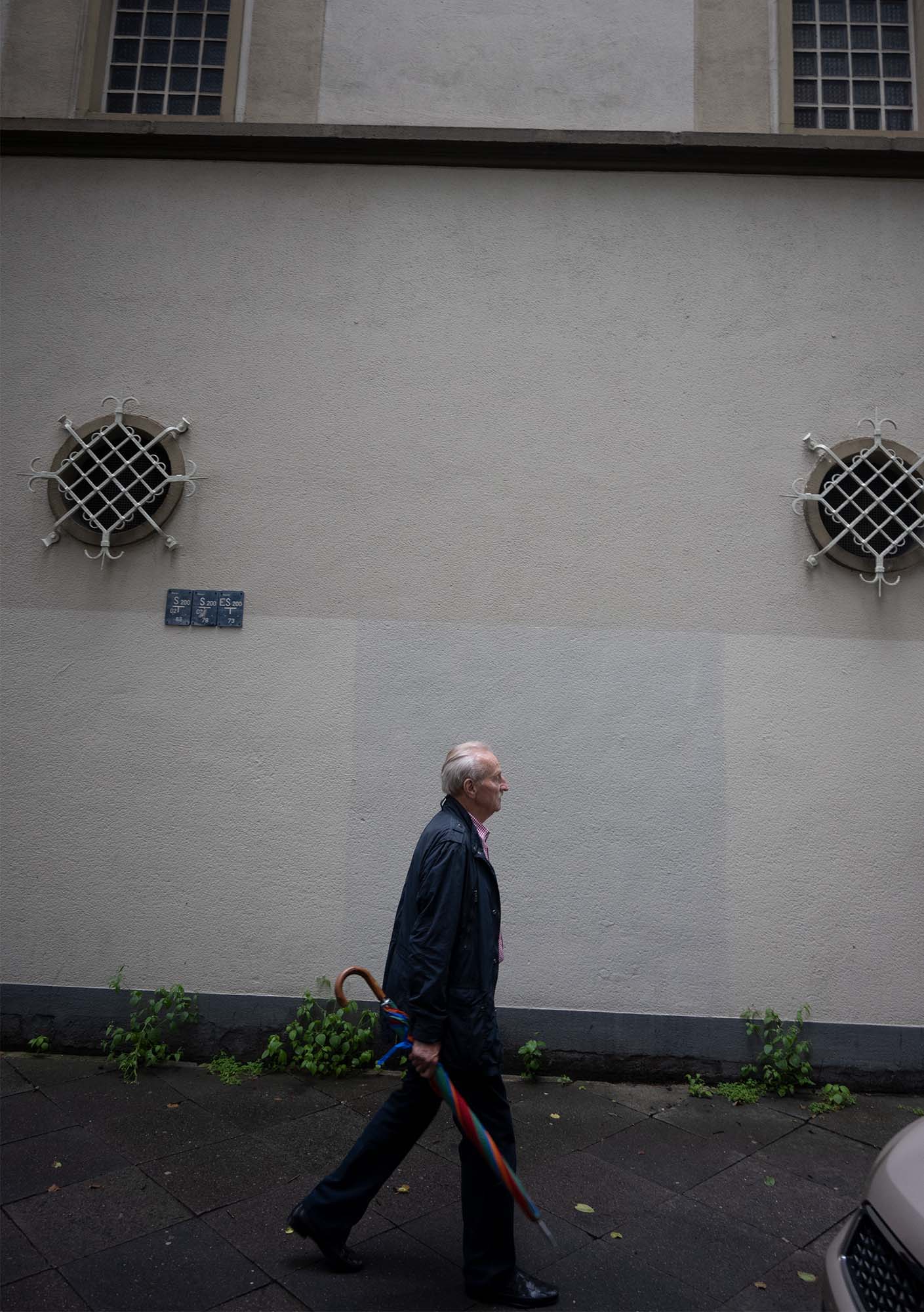 Die Katze läuft mit einem geschlossenen Regenschirm in der Hand vor einer Fassade entlang