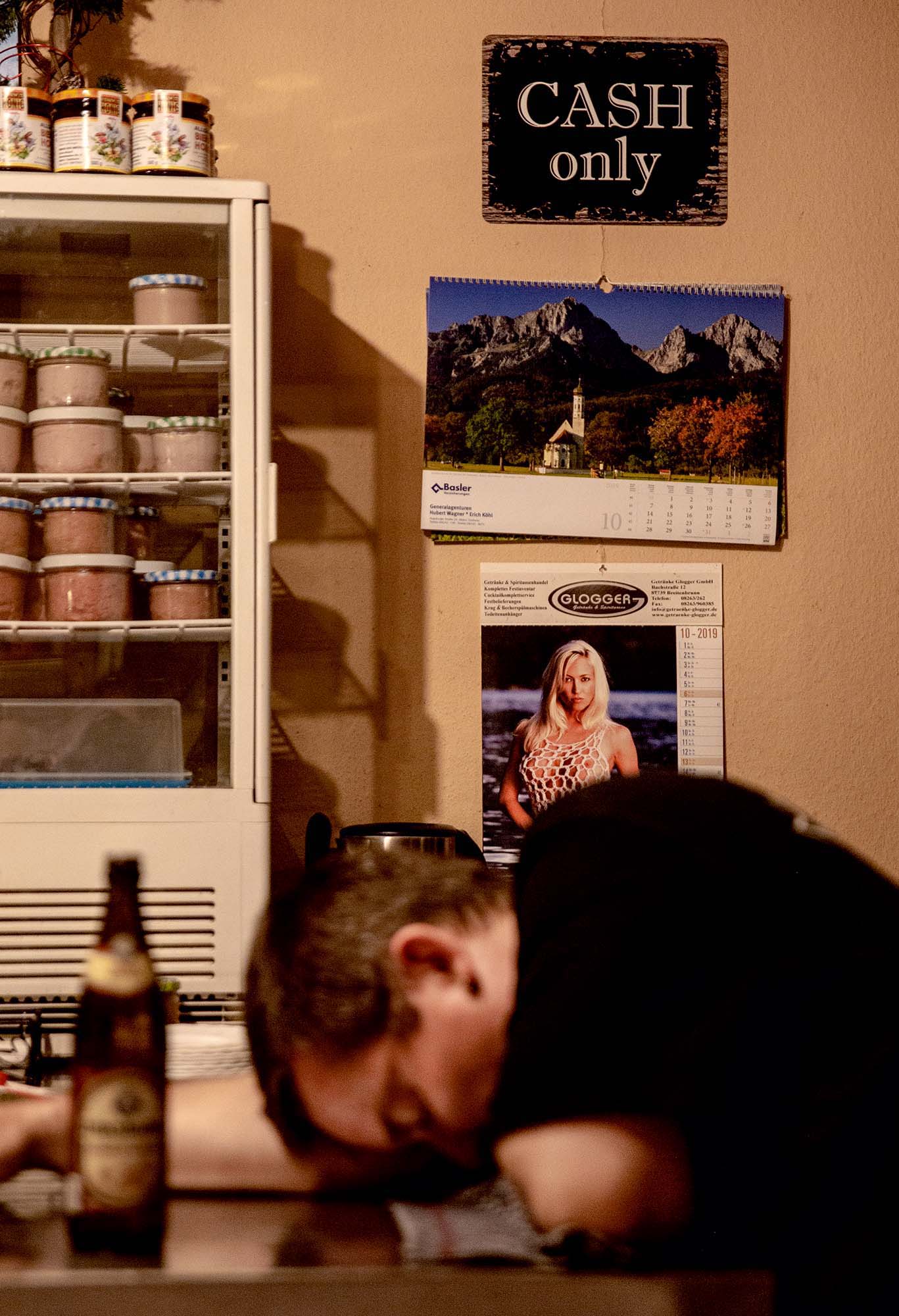 Ein Mann schläft auf dem Tresen. Hinter ihm an einer Wand hängen ein Schild mit der Aufschrift: Cash only, ein Kalender mit einem Bild einer Kirche vor hohen Bergen und ein zweiter Kalender mit einer megatitten Blondine