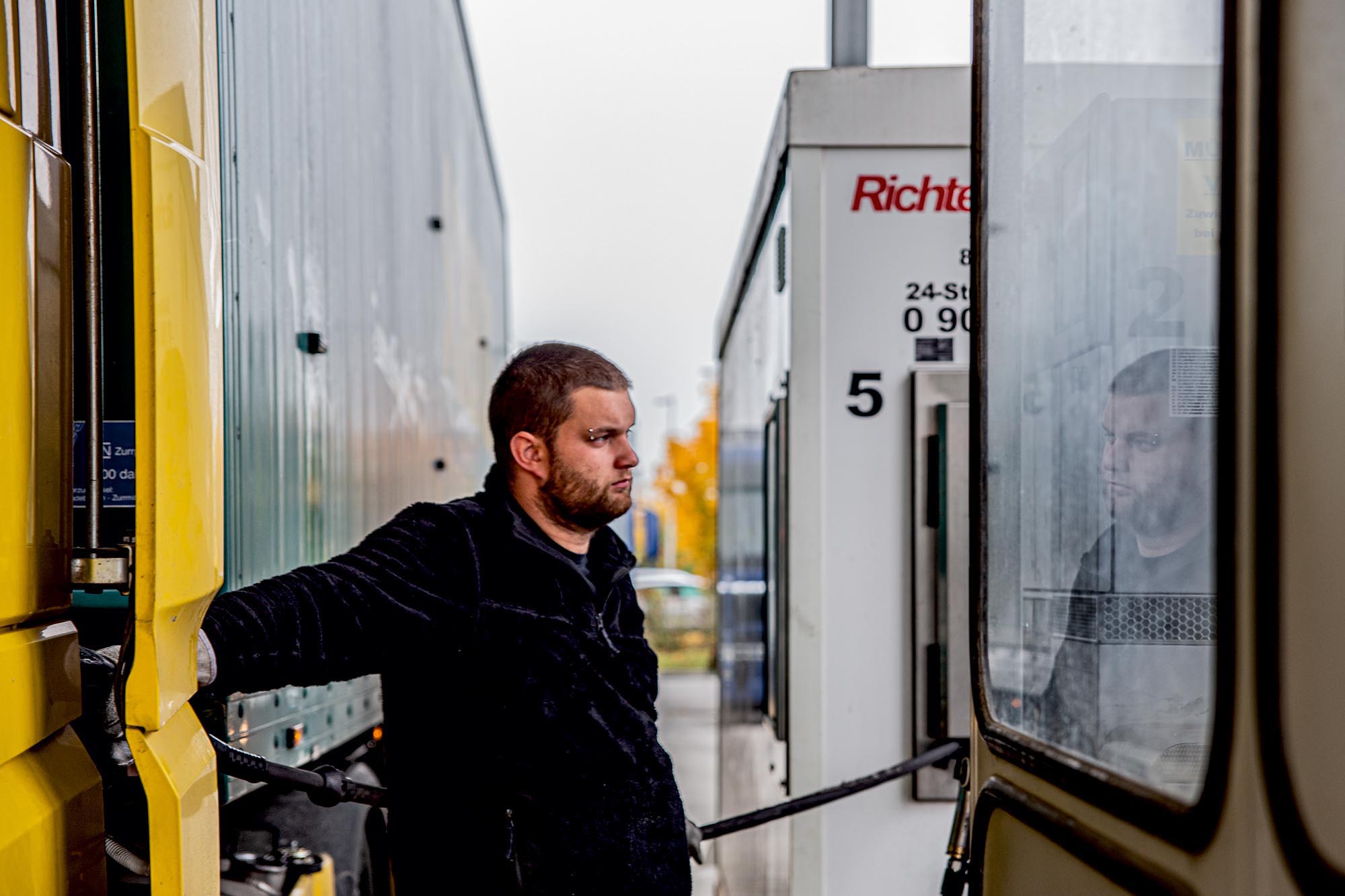 Michl steht an einer Zapfsäule. Sein ernster Blick spiegelt sich in einem Fenster